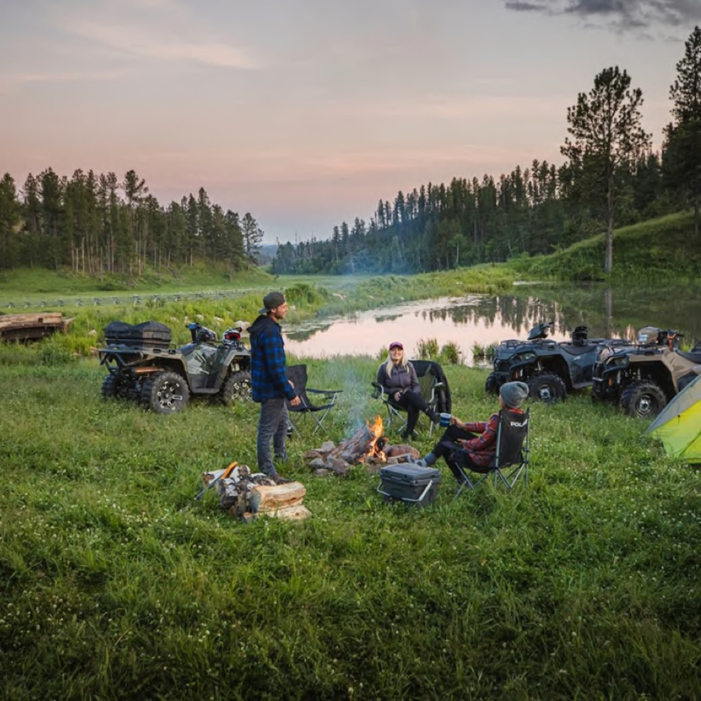 A small group of people gathered around a campfire with several all-terrain vehicles (ATVs) nearby. They are in a grassy campground near a calm pond, with trees in the background and a pastel sunset sky.