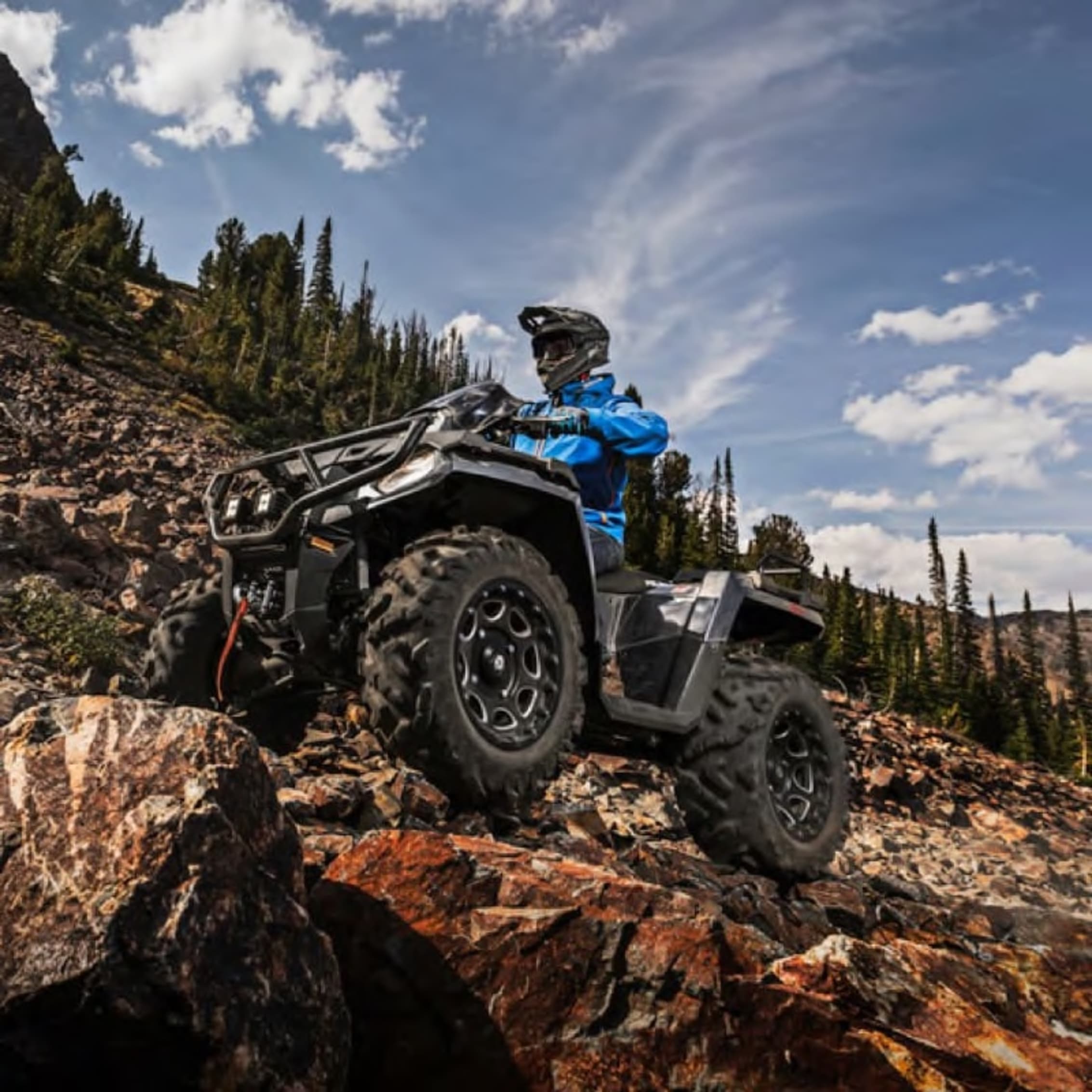 A person riding an all-terrain vehicle (ATV) on rocky terrain in a mountainous park, surrounded by tall trees and a bright blue sky.
