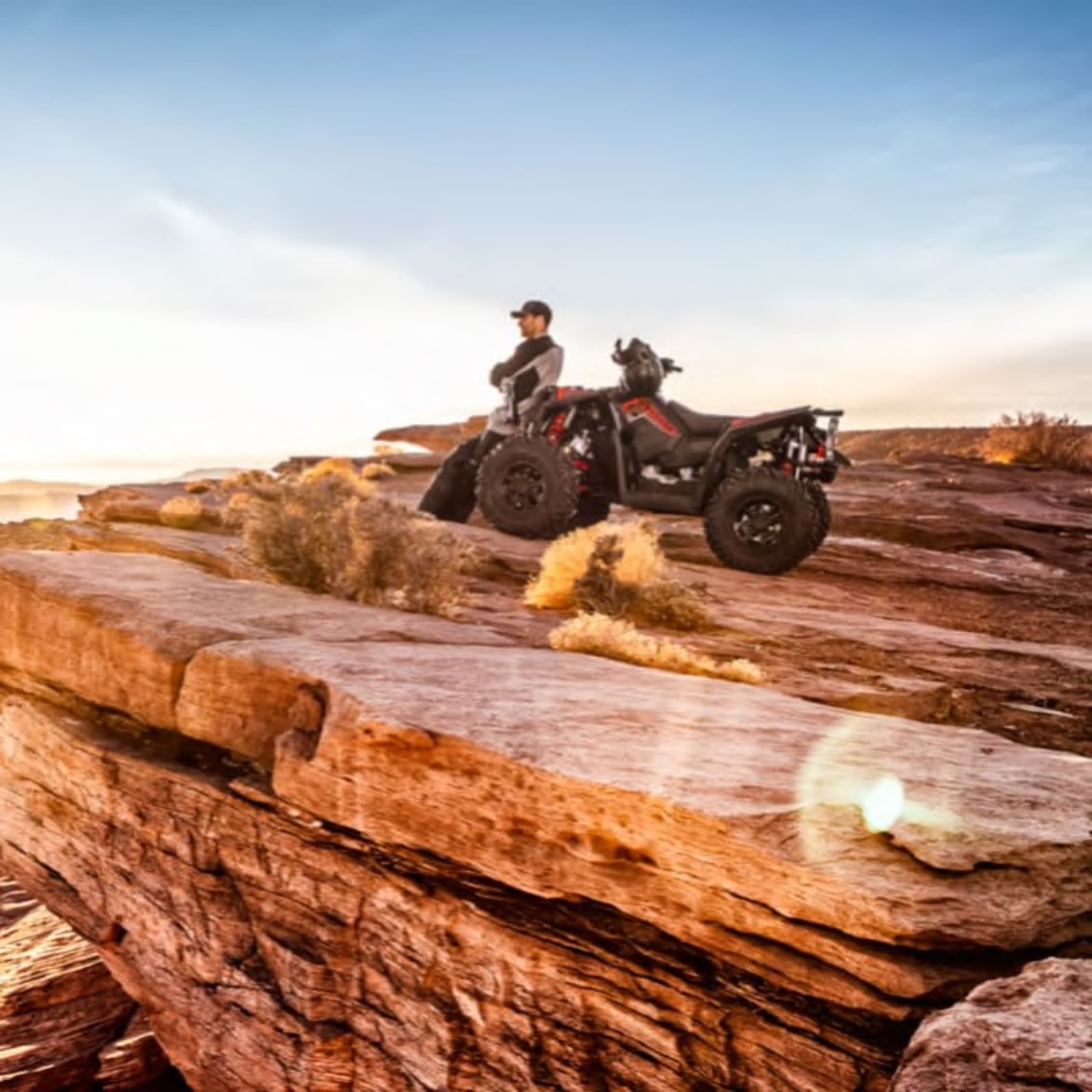 A person standing beside an ATV on a rocky cliff, gazing out at a scenic outdoor landscape under a bright sky.