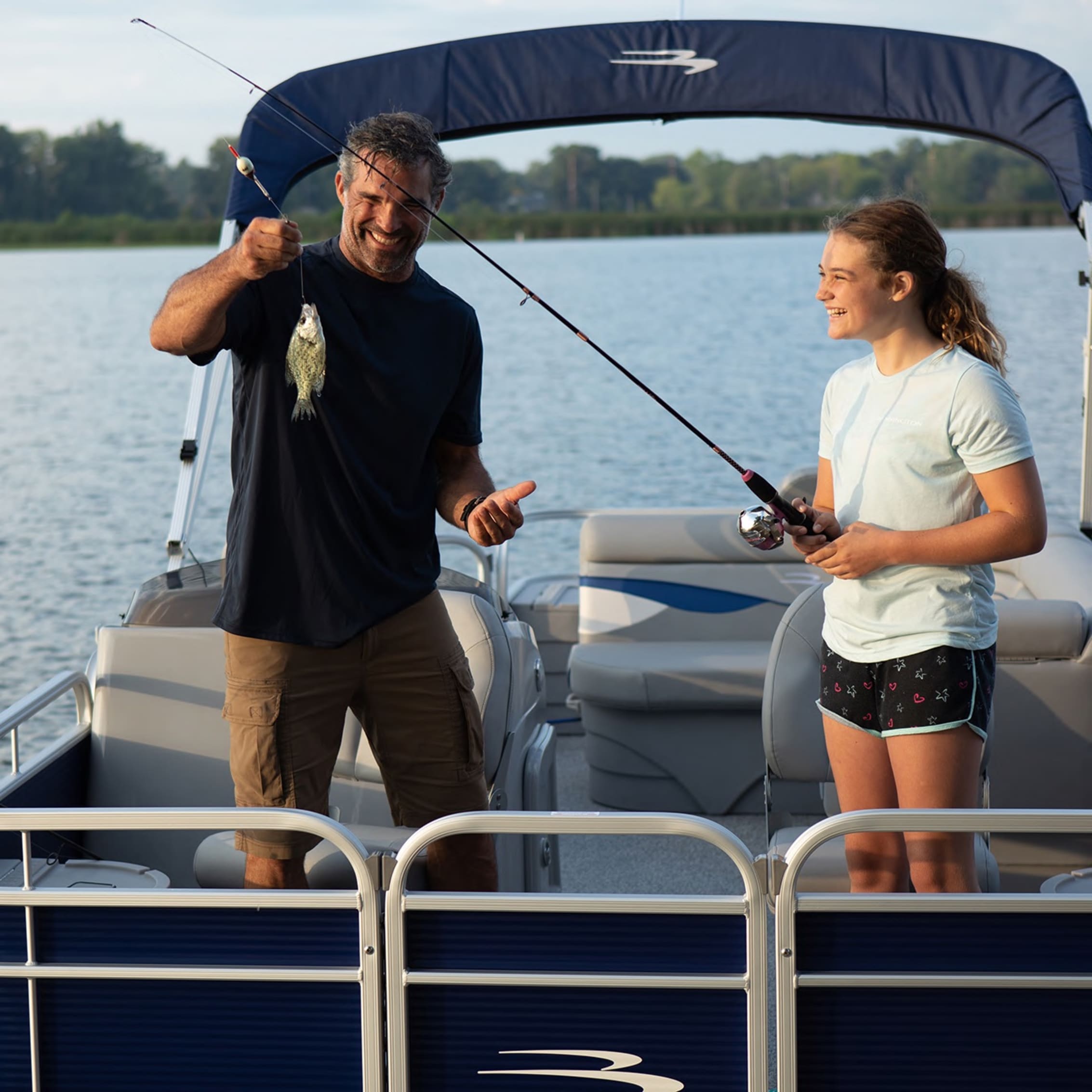 A man and a young girl fishing on a pontoon boat, with the man holding up his catch and both smiling against a scenic lake backdrop.