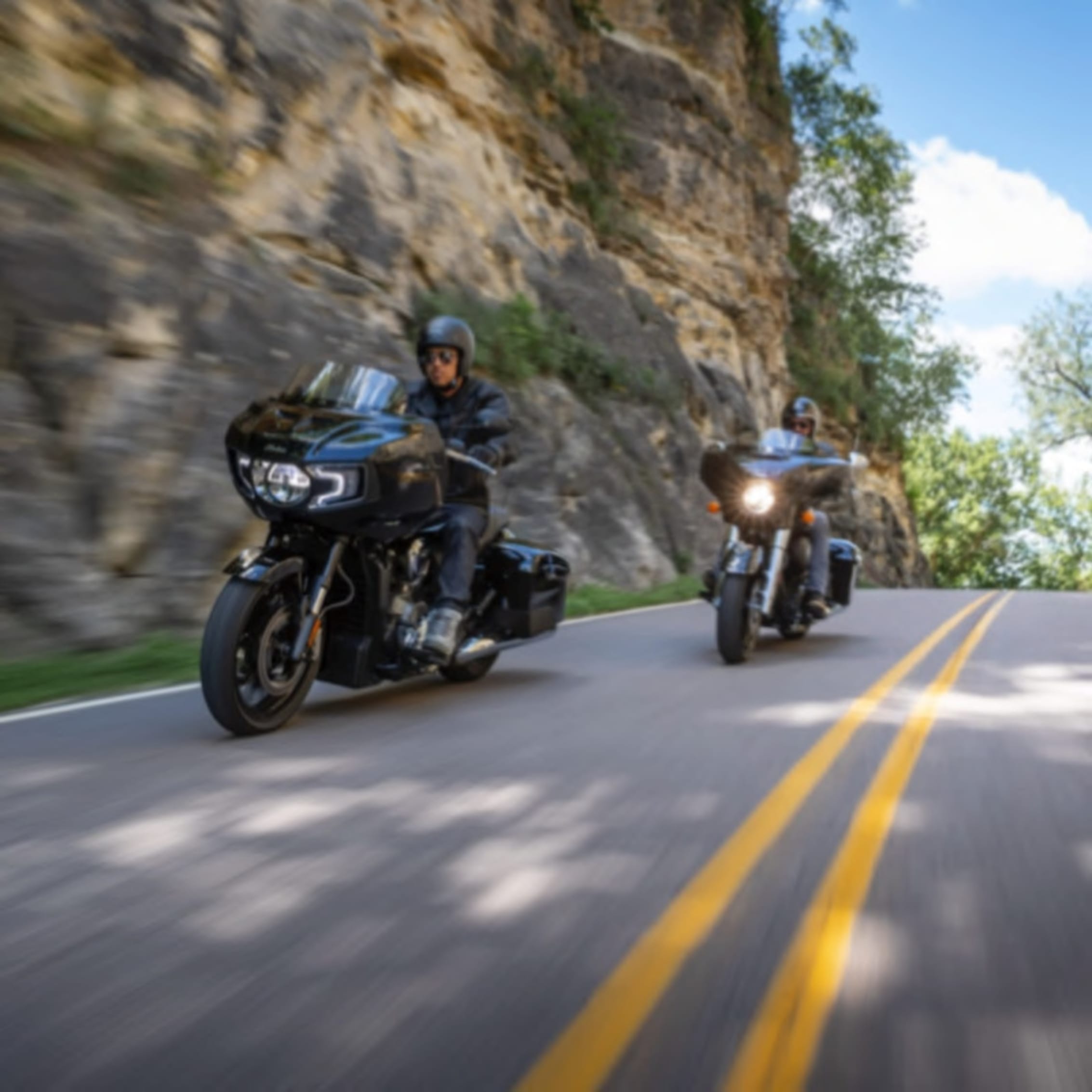 Two motorcyclists riding dynamically on a winding road beside a rocky cliff, highlighting the exhilaration of a ride in the great outdoors.