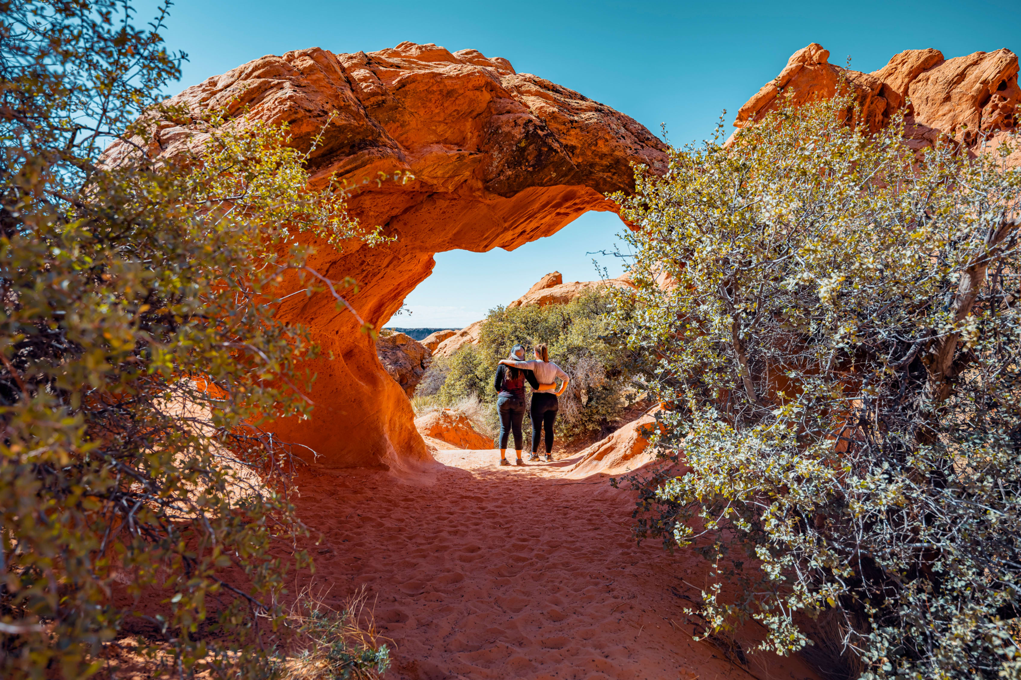 Two women stand on a sandy trail looking through a large, natural red rock arch.