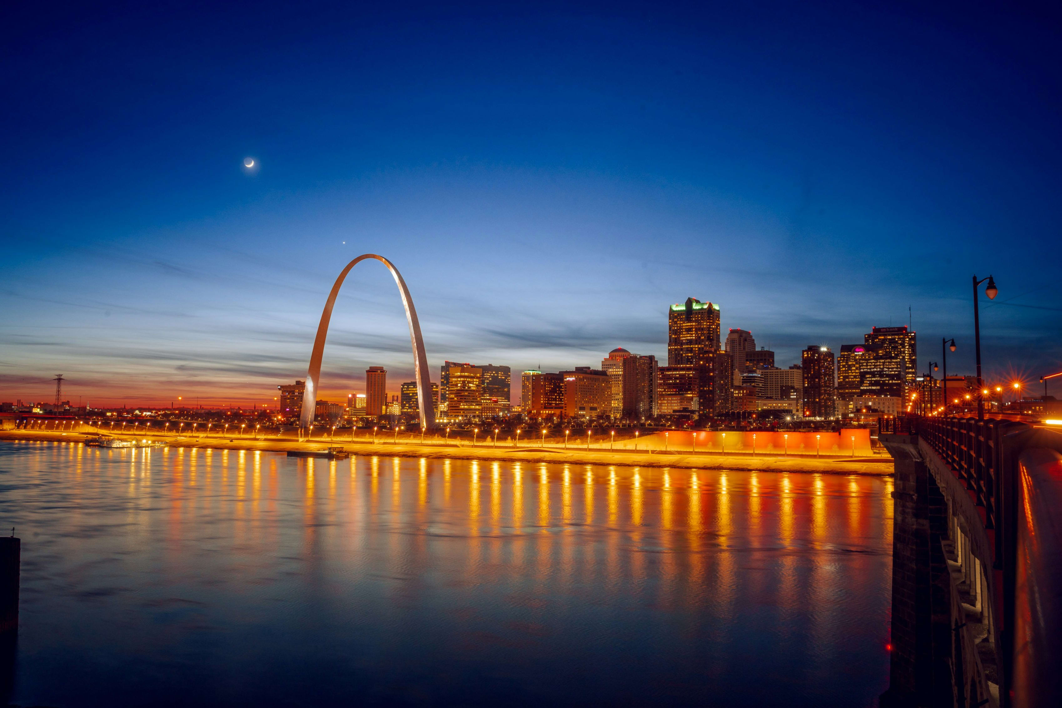 Dusk view of Gateway Arch reflecting on water with city skyline and bridge in background