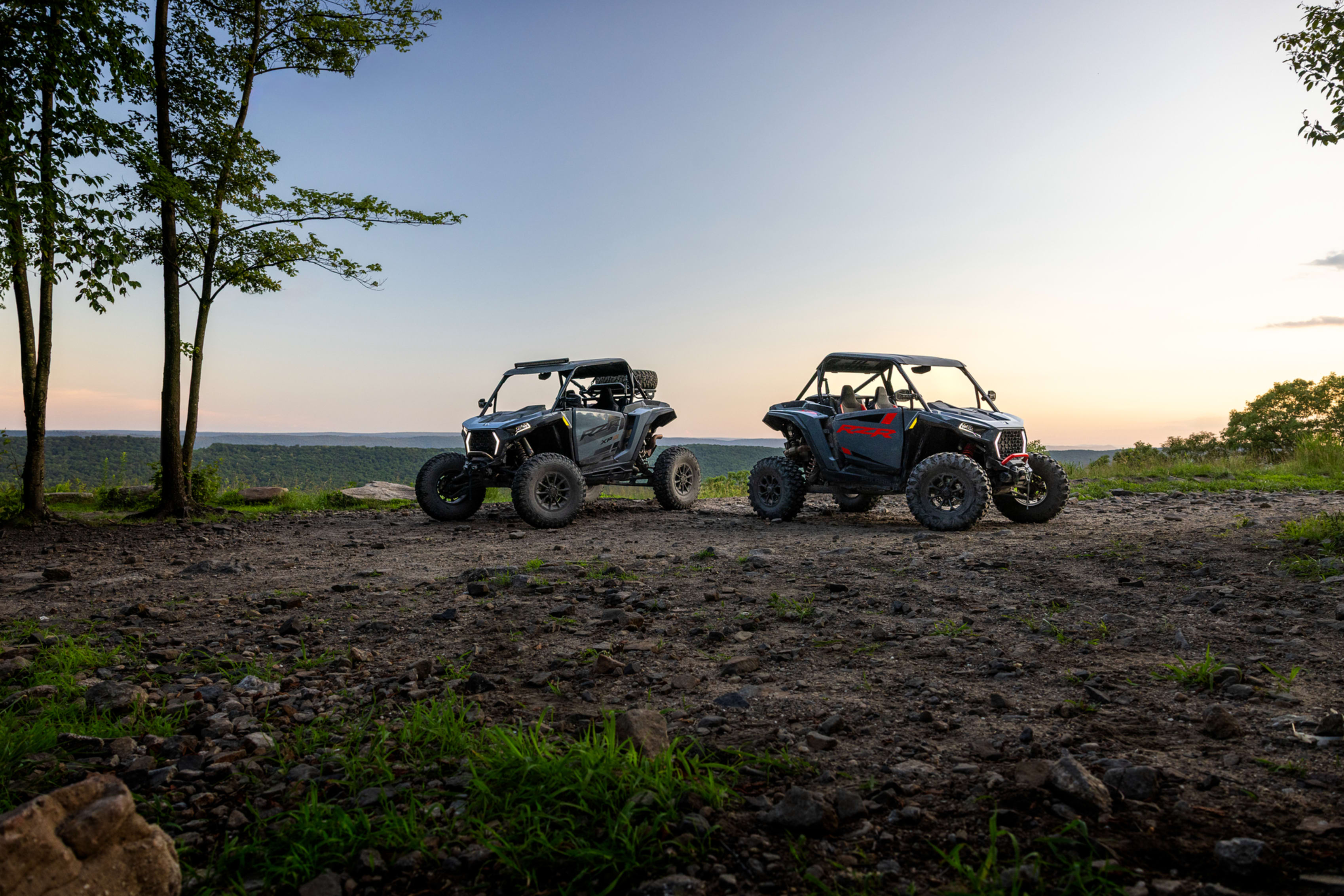 Two Polaris RZRs parked on a rocky trail at sunset with hills in the background.