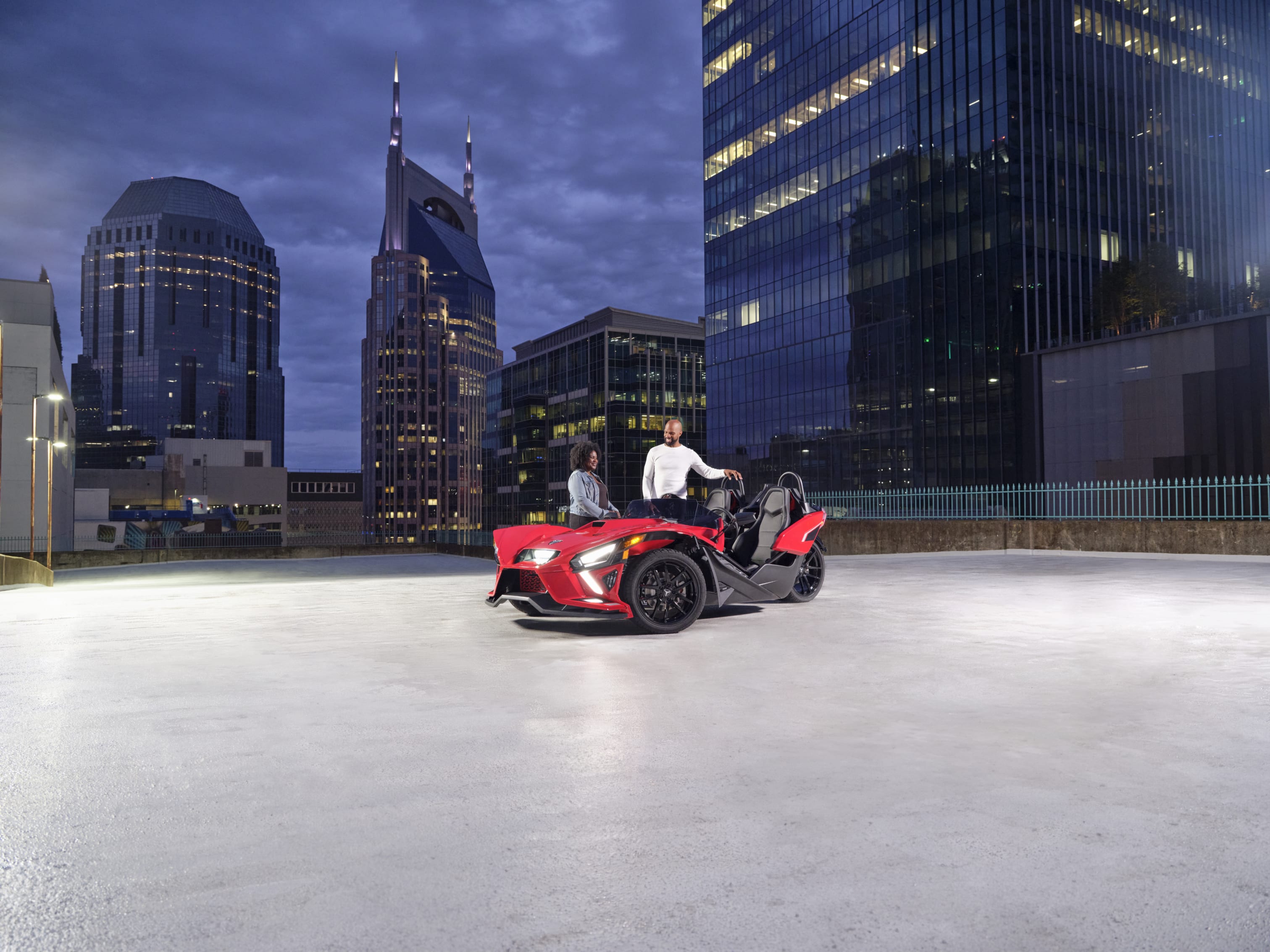 Couple enjoying an evening together with a red Polaris Slingshot against a backdrop of city skyscrapers at dusk.