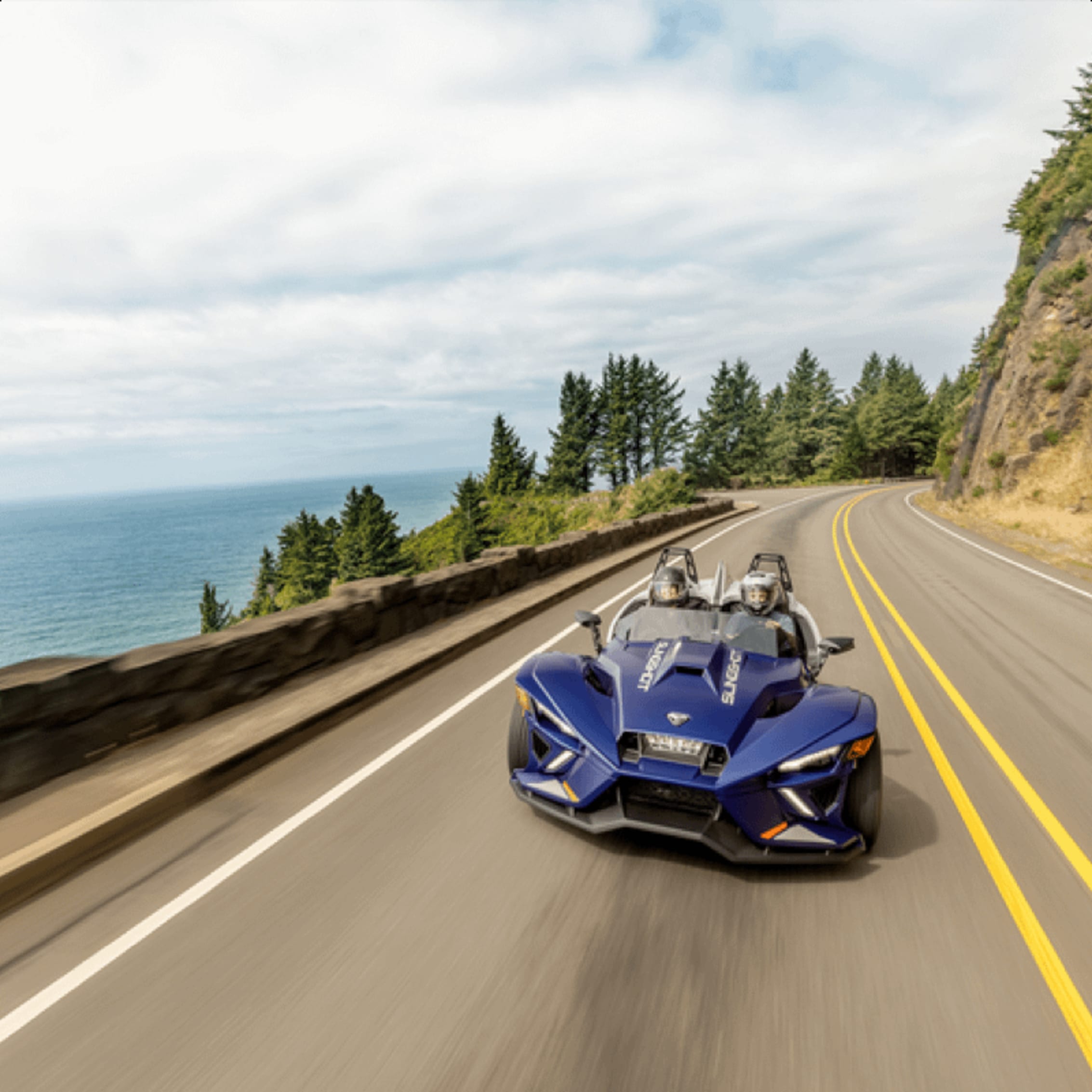 A blue Polaris Slingshot cruising along a winding road by the ocean, with a scenic view of trees and cliffs in the background.