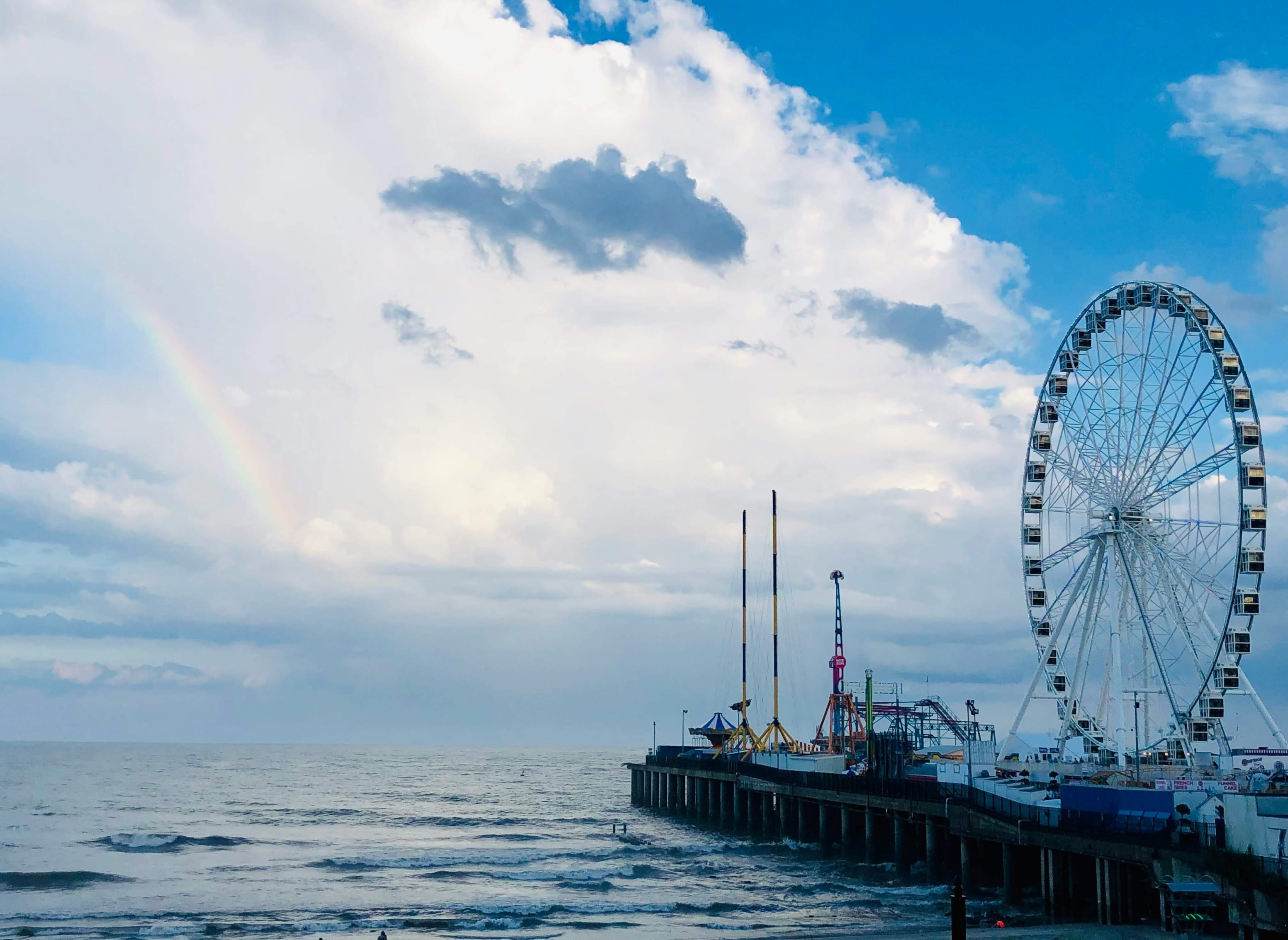 New Jersey pier with a large Ferris wheel and amusement rides overlooking the ocean under a sky with a faint rainbow.