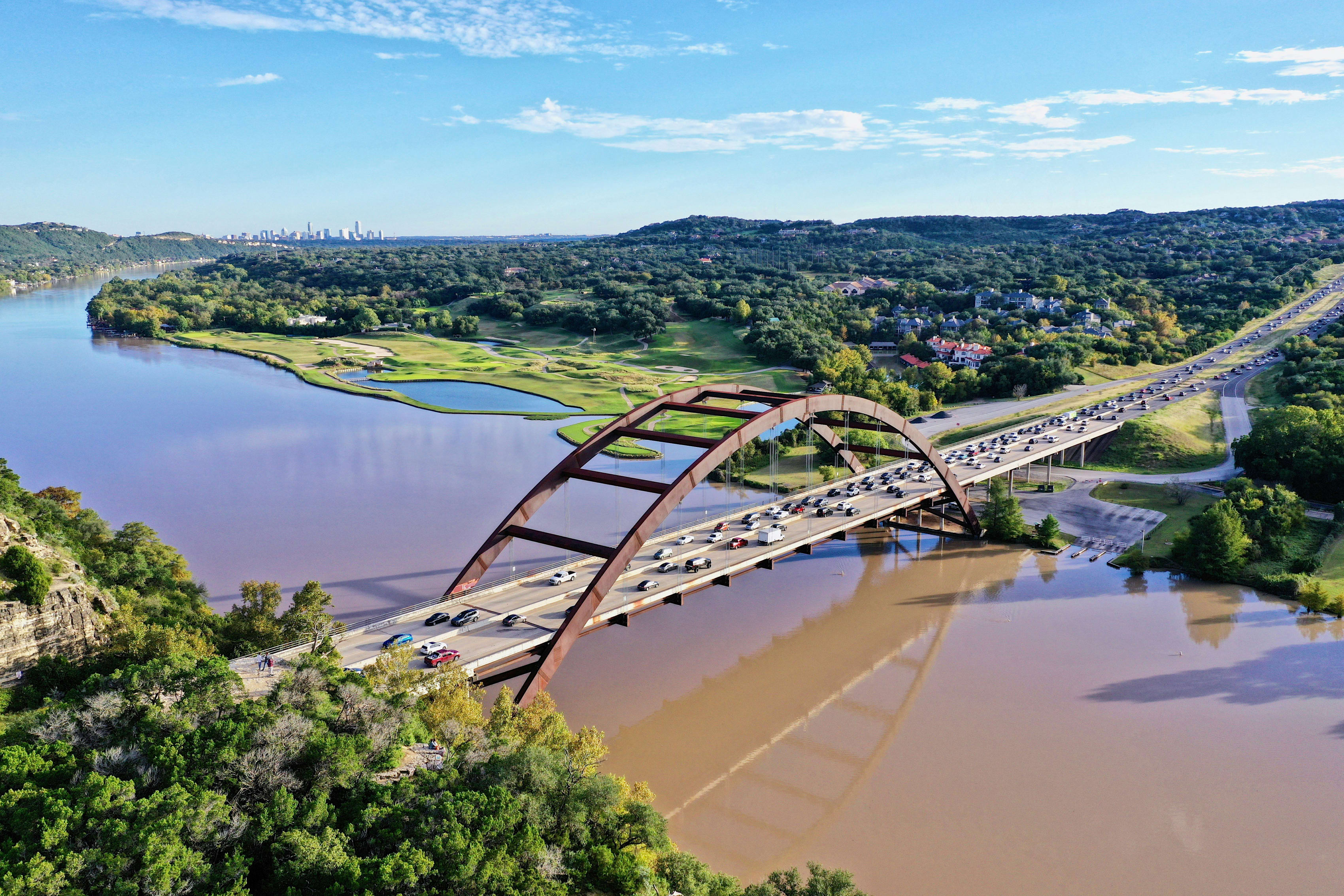 Aerial view of a distinctive arched bridge over a river in Texas with lush greenery and city skyline in the distance.