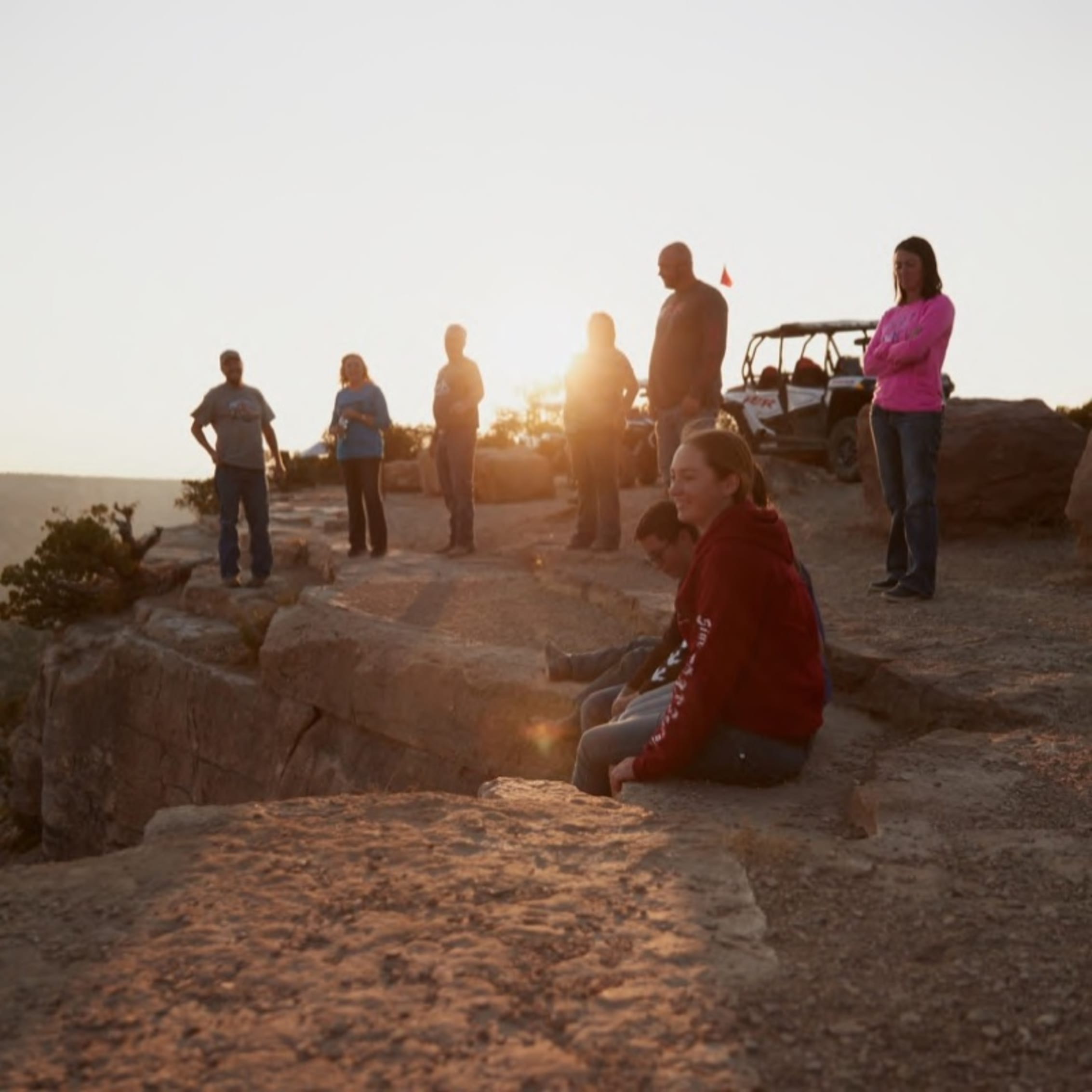A group of people at the edge of a rocky overlook during sunset, with some seated and others standing, capturing a scenic outdoor adventure.