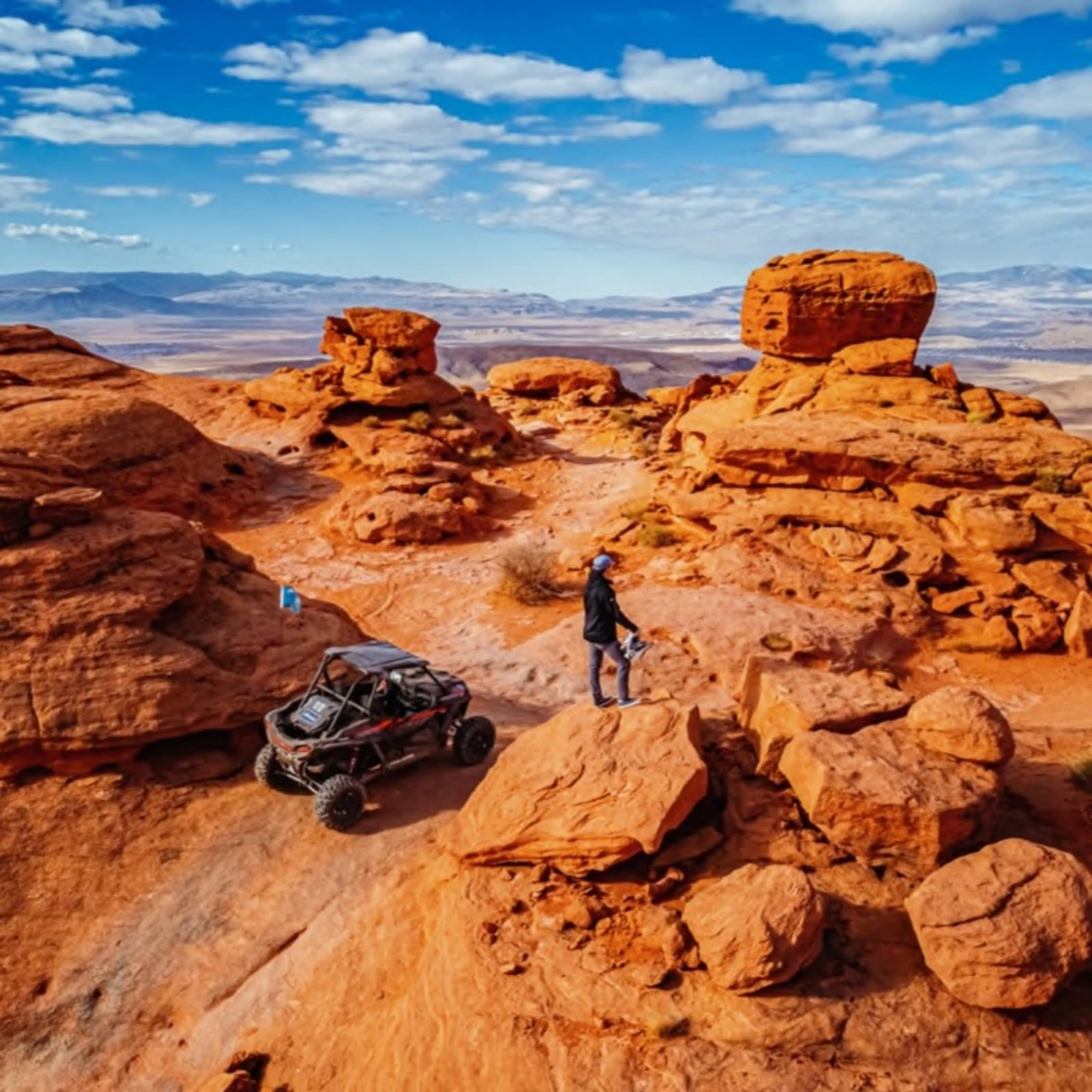 A person standing on a large rock formation in a desert landscape, with a four-wheeler nearby, against a backdrop of stunning red rock formations under a blue sky with scattered clouds.