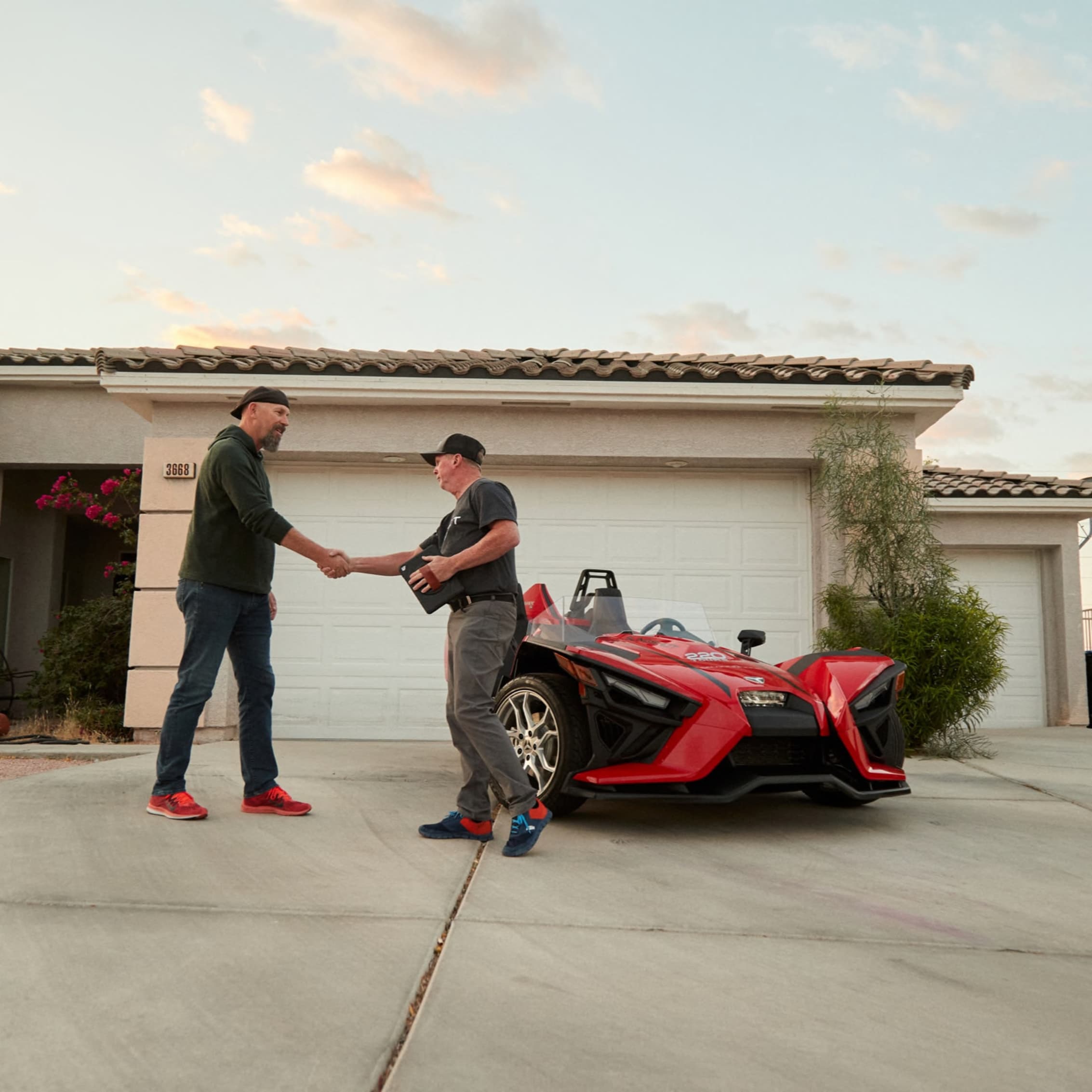 Two men shaking hands in a driveway, with one about to hand over the keys to a red Polaris Slingshot, indicating a personal touch in vehicle pickup and delivery service.