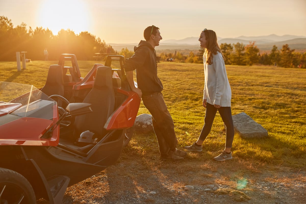 Couple talking beside a Polaris Slingshot in a field at sunset.