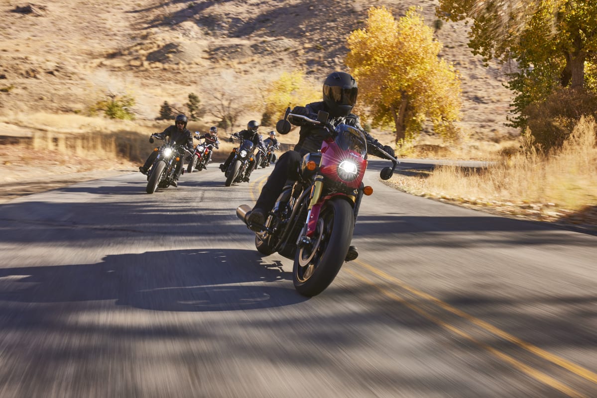Group of riders on Indian motorcycles cruising on a sunlit road with autumn trees in the background.