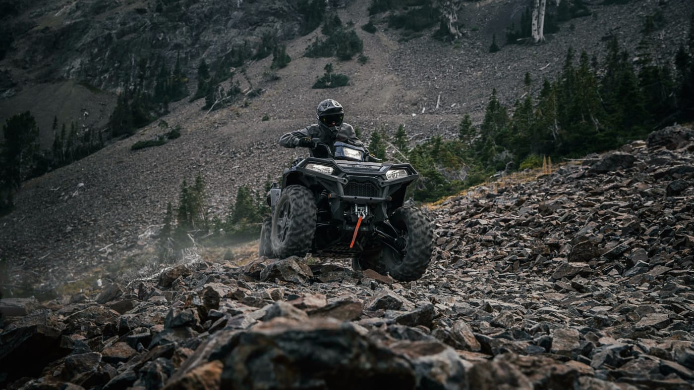 A rider wearing a full-face helmet is off-roading on an all-terrain vehicle (ATV) over a steep, rocky terrain in a forested mountainous area.