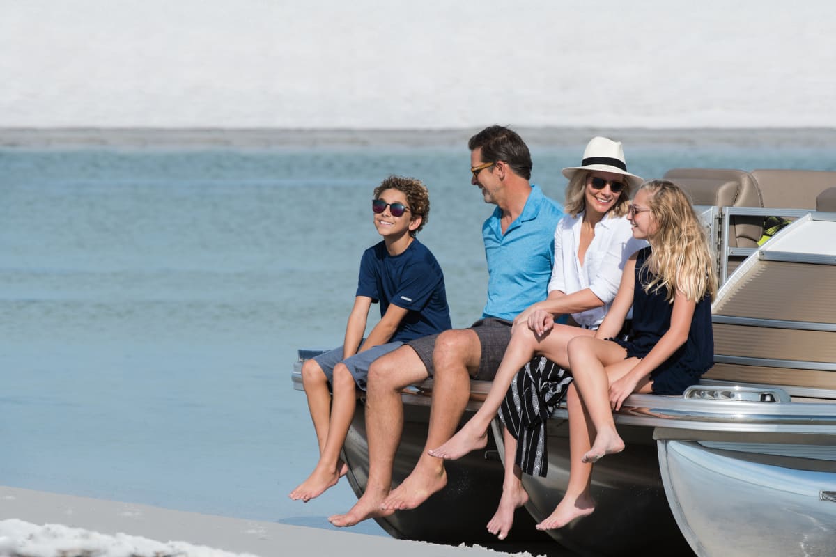 Family enjoying time together sitting on the edge of a Bennington pontoon with legs dangling over a sandy beach.