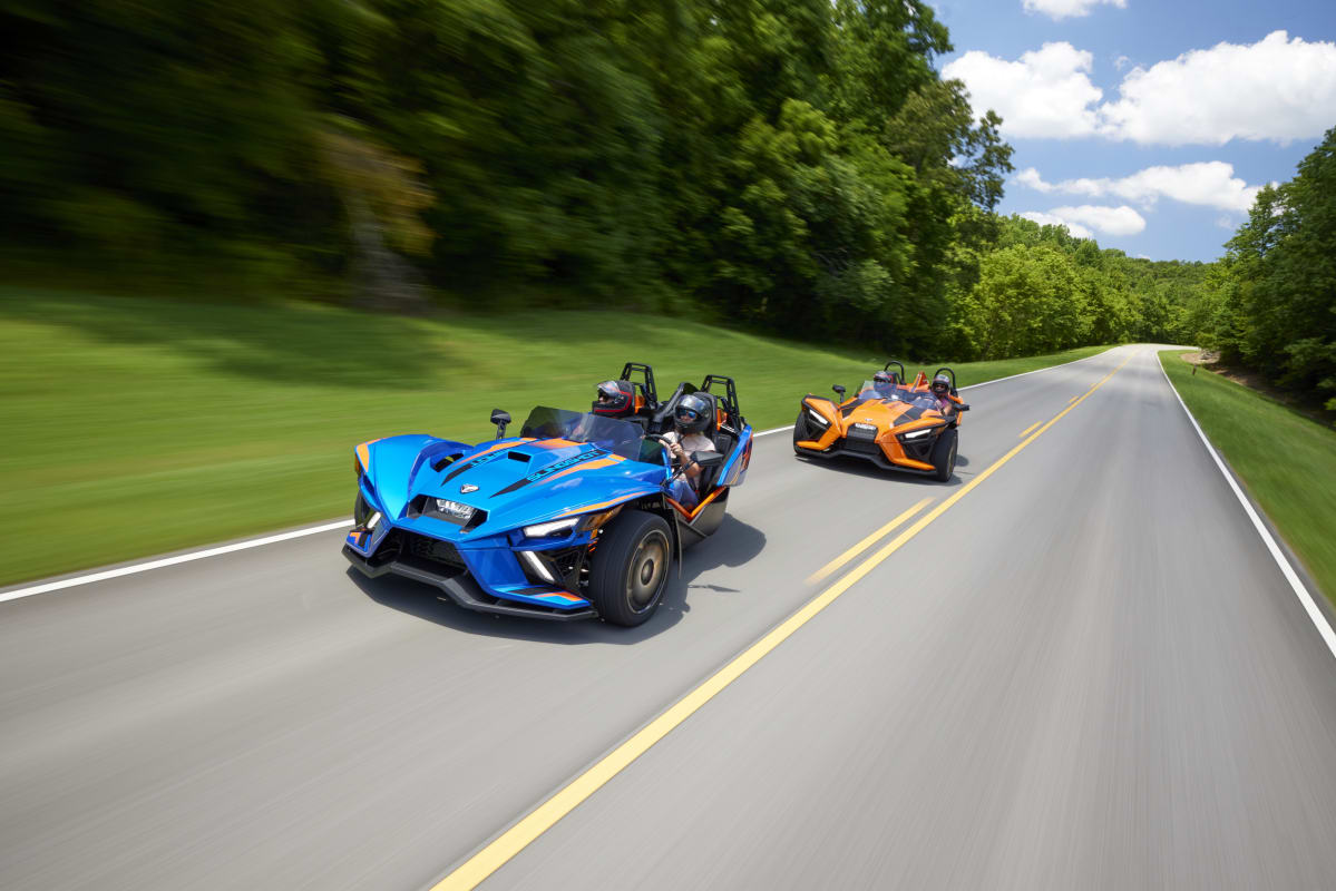 A blue and an orange Polaris Slingshot driving down a scenic tree-lined road on a sunny day.