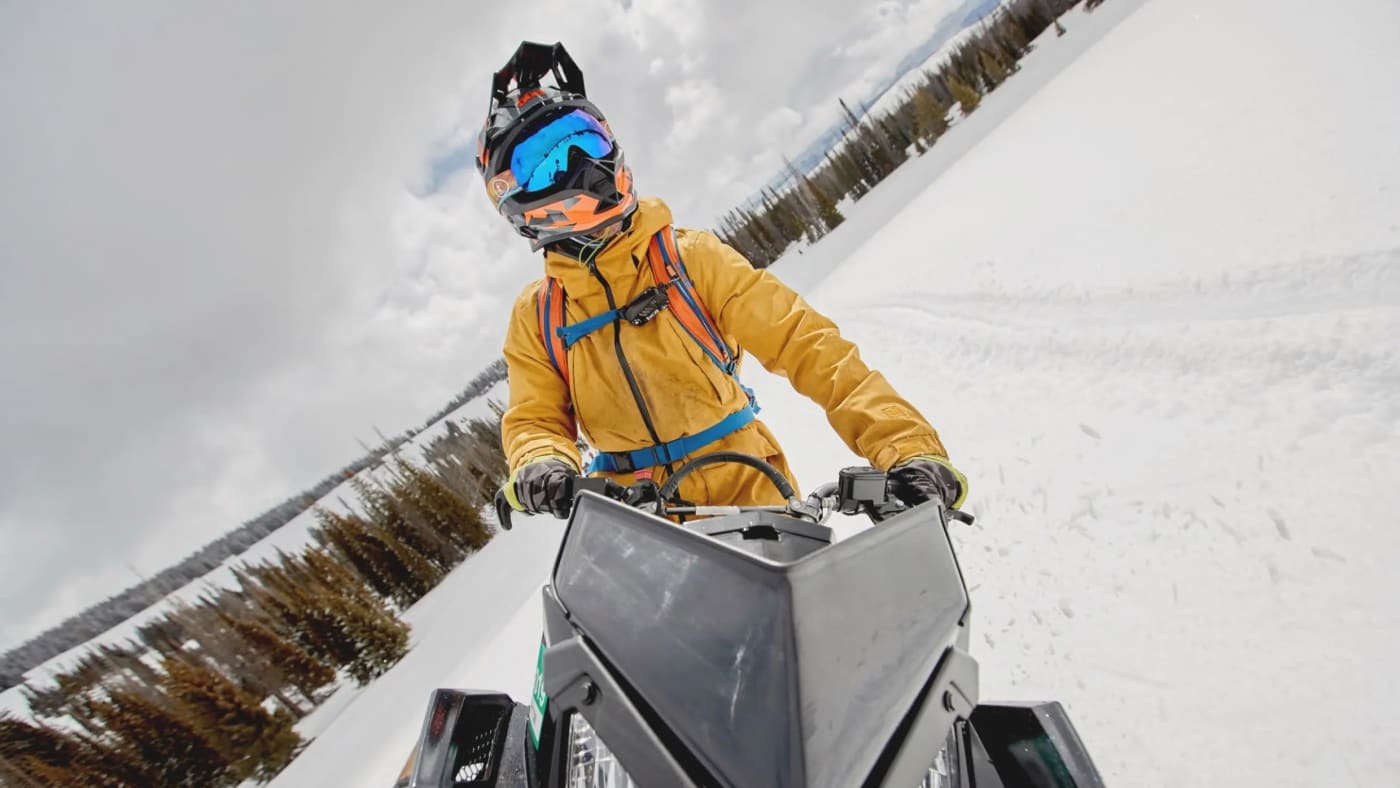 A person in a bright yellow jacket and helmet rides a snowmobile through a snow-covered landscape with trees in the background.
