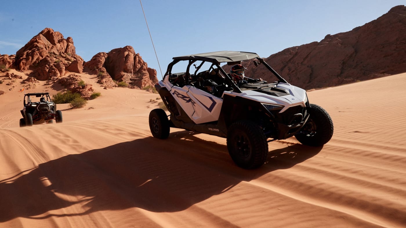 Two side-by-side off-road vehicles driving through a sandy desert with rocky formations in the background.