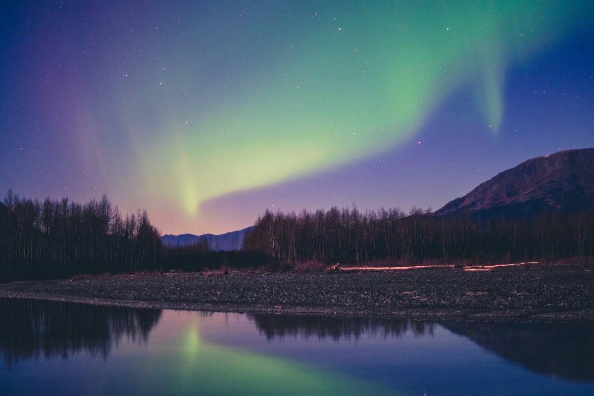 Aurora borealis over an Alaskan river with reflection and forest silhouette at twilight.