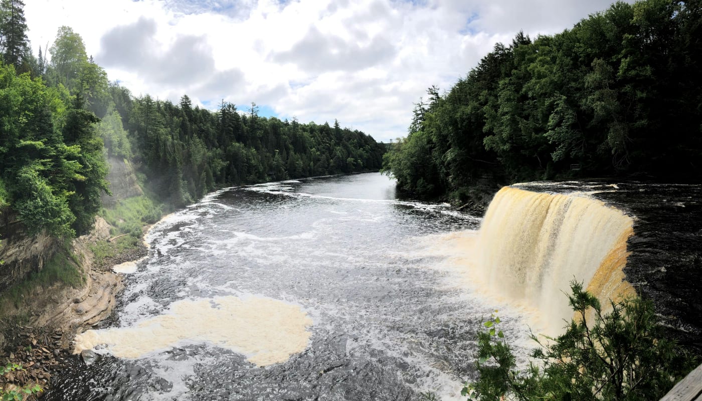 Panoramic view of a large waterfall in Michigan surrounded by lush forests on a cloudy day.