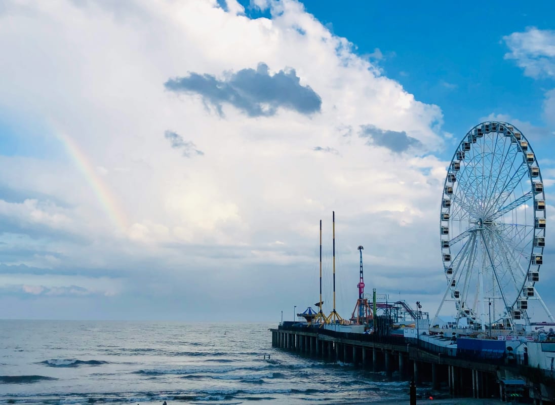 New Jersey pier with a large Ferris wheel and amusement rides overlooking the ocean under a sky with a faint rainbow.