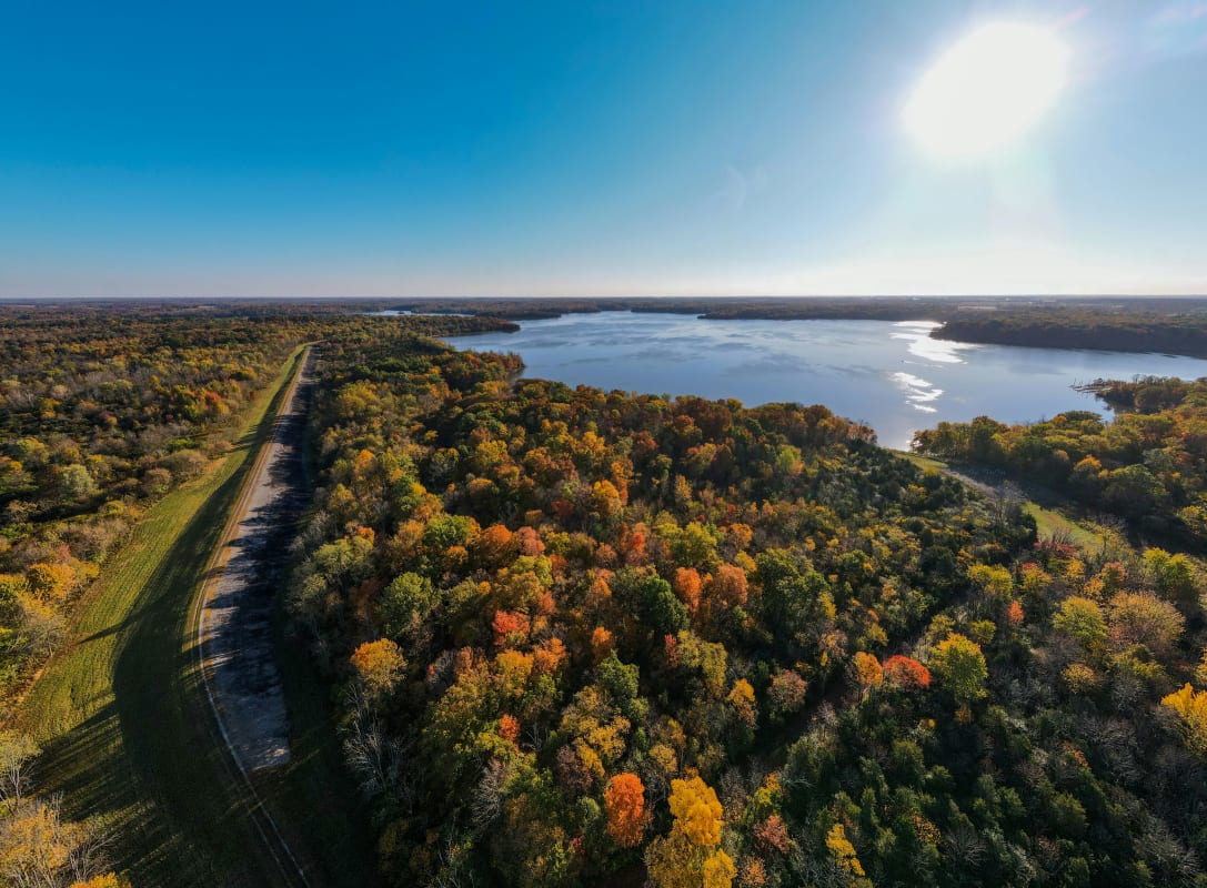 Aerial view of Ohio landscape with a mix of dense autumn-colored trees and a sparkling lake under a clear blue sky.