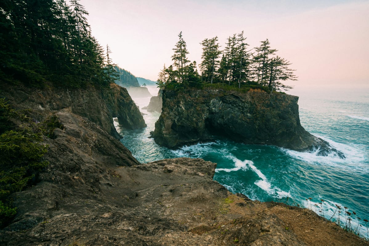 Scenic view of Oregon coastline with waves crashing against rugged cliffs and evergreen trees at dusk.