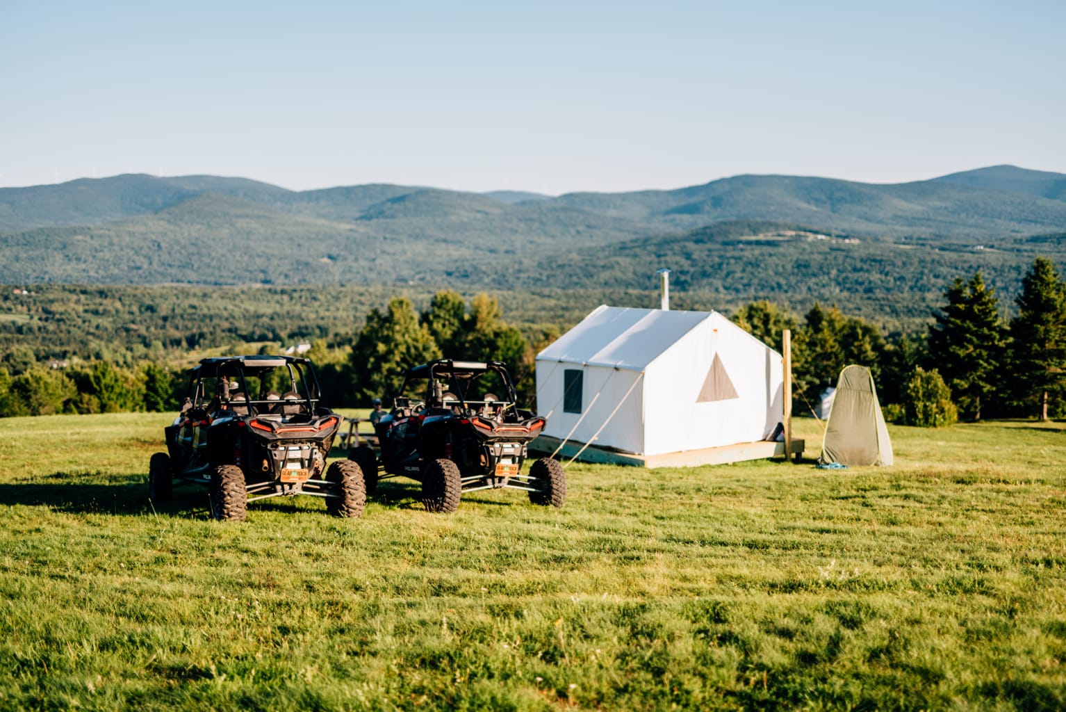 Two Polaris RZRs parked beside a canvas tent overlooking rolling mountains