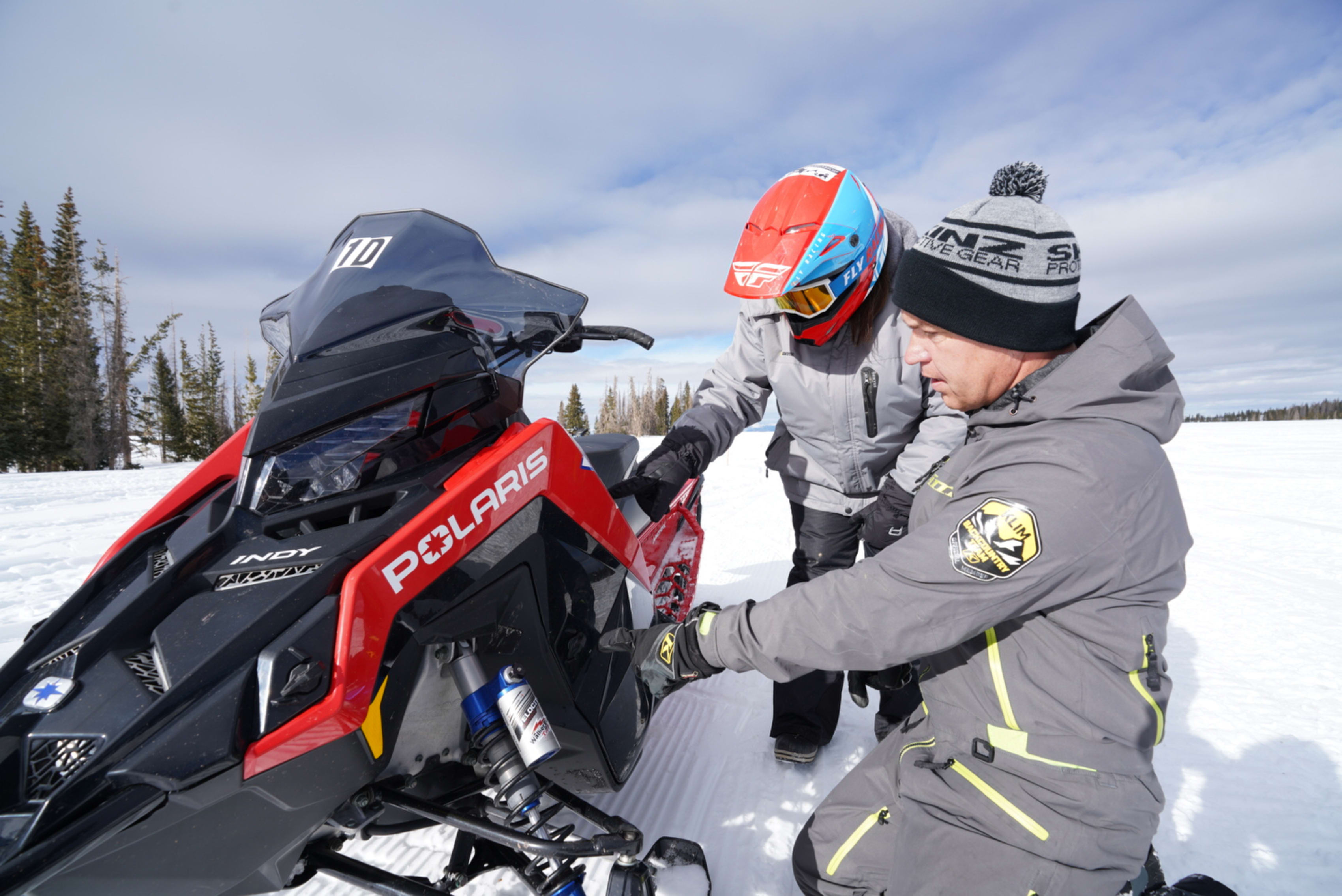 Two individuals in winter gear inspecting a Polaris snowmobile on a snowy terrain under clear skies.