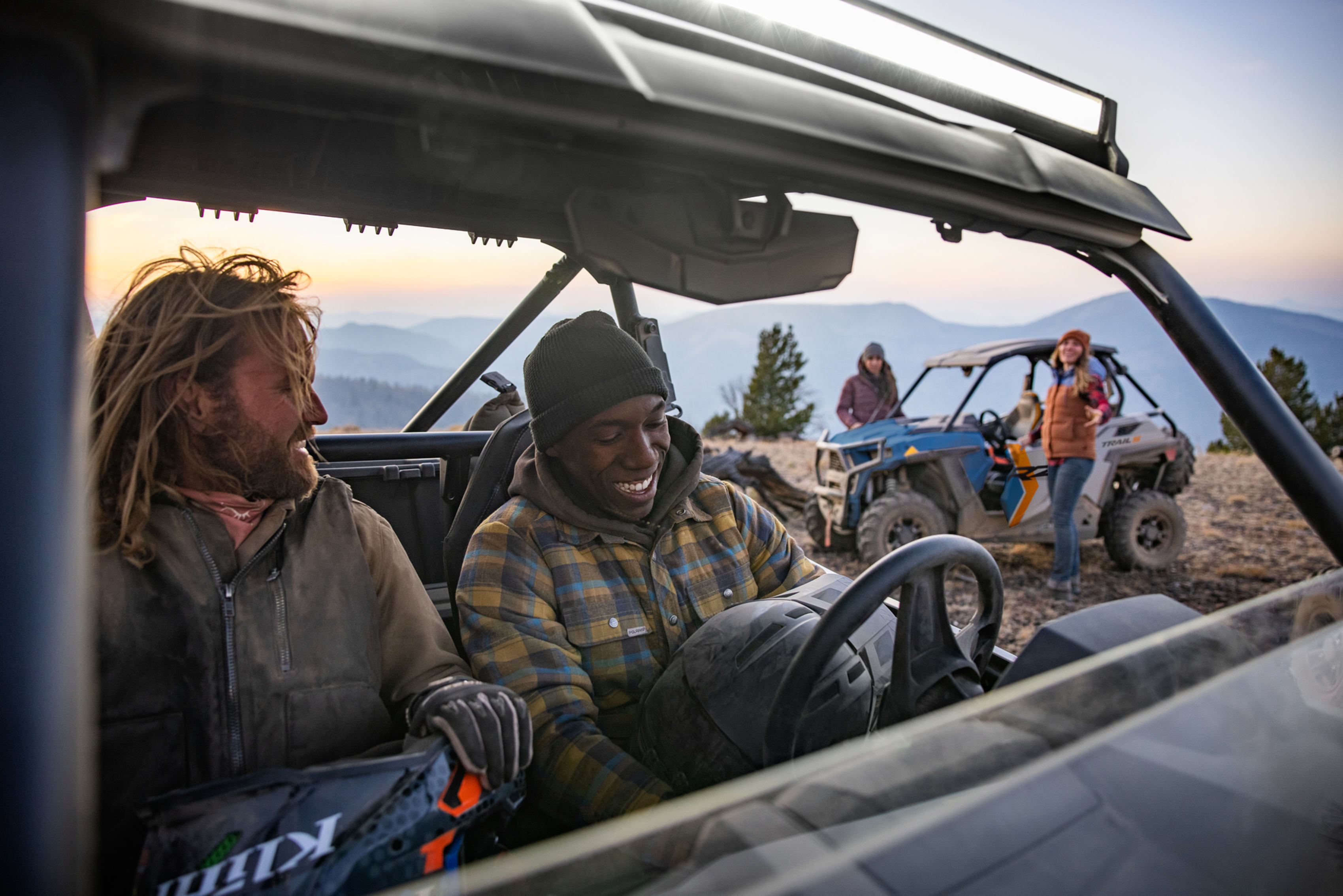Joyful adventurers sharing a laugh in a Polaris off-road vehicle during a mountainous sunset.