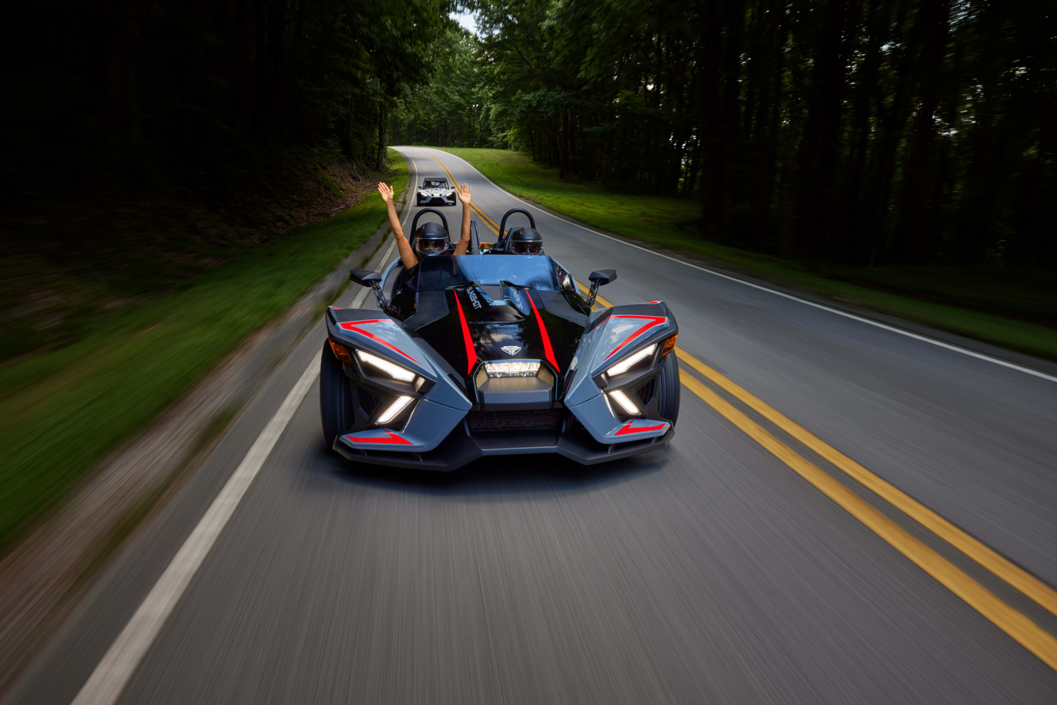 Exhilarating ride in a Polaris Slingshot on a tree-lined road with passenger expressing joy.