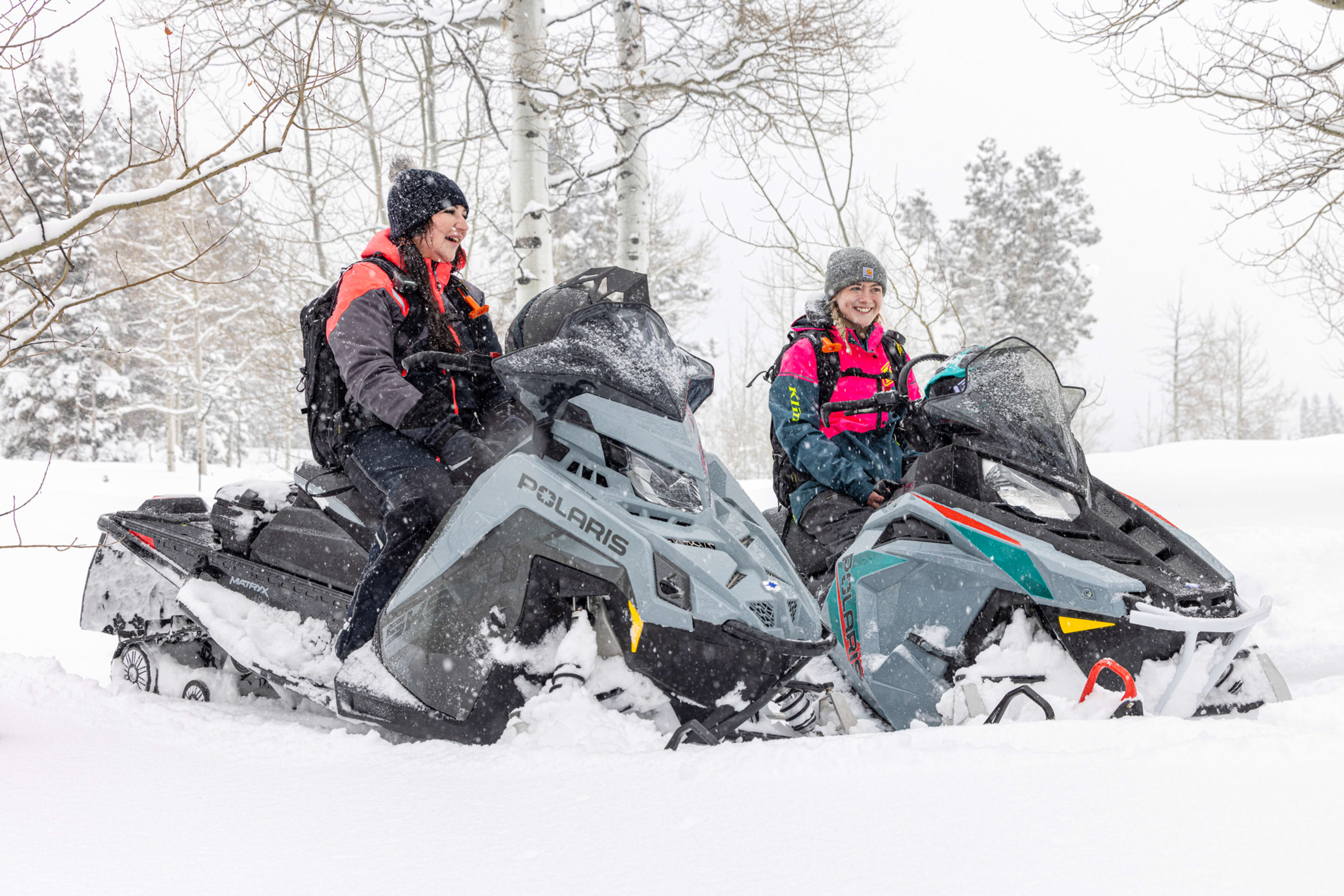 Two people joyfully riding snowmobiles through a snowy forest, with snowflakes falling around them and trees lightly dusted with snow in the background.