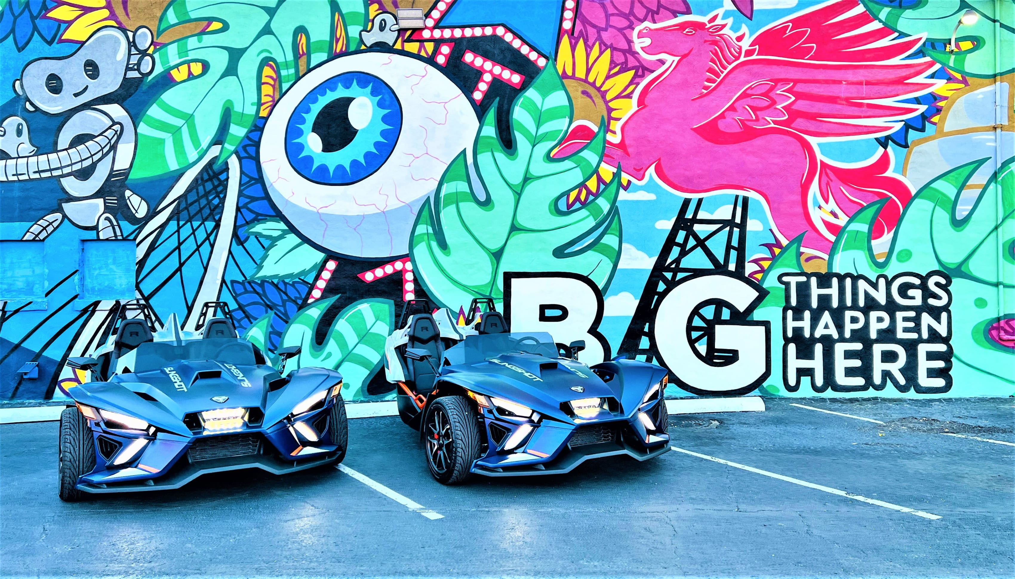 Two blue Polaris Slingshots in front of art wall